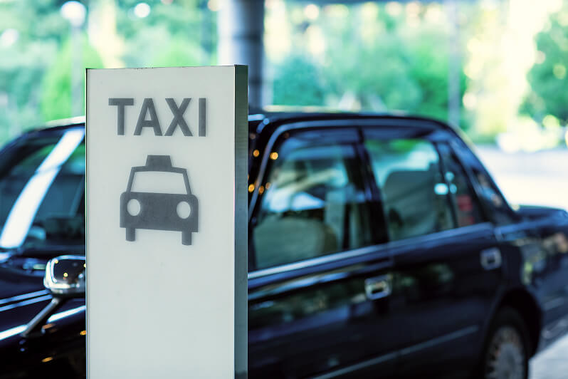 「TAXI」という文字と車のアイコンが描かれた縦型の看板と、その奥に停車している黒いタクシーの車体。